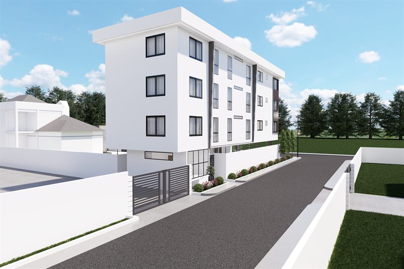 The block from the road at an angle, the flank running away with windows stacked over four storeys, balconies on the far corner, and a slatted gate set in the white boundary wall with clipped shrubs along it.